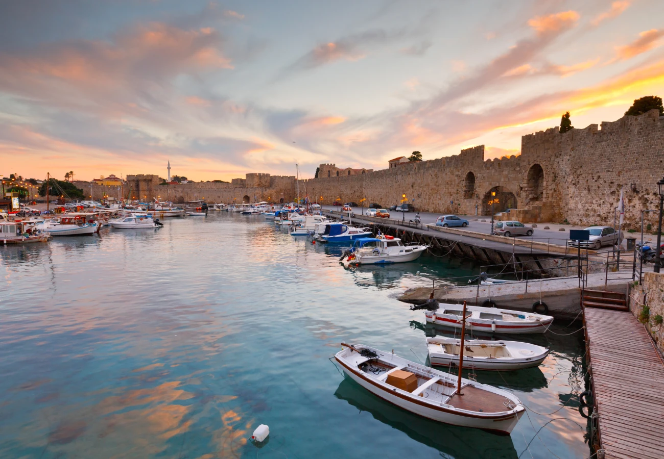 Scenic view of Rhodes harbor at sunset, featuring moored boats and medieval stone walls along the waterfront.