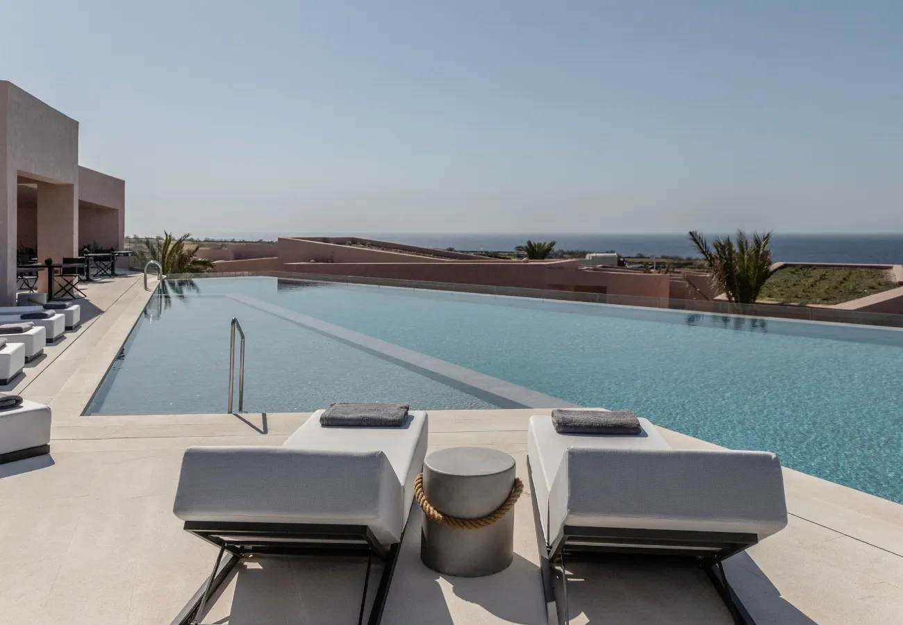 Modern outdoor pool terrace with sun loungers overlooking the sea under a clear blue sky, at Domes Novos Santorini luxury resort