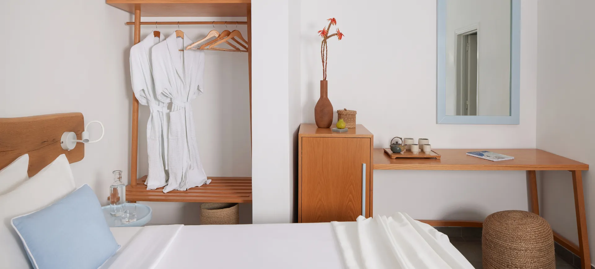 Contemporary guest room in Santorini with white walls, natural wood furniture, and soft blue accents. A neatly made bed sits beside a small table with a glass water carafe, while an open wardrobe displays two white bathrobes. A wooden desk with a mirror, tea set, and woven stool completes the space
